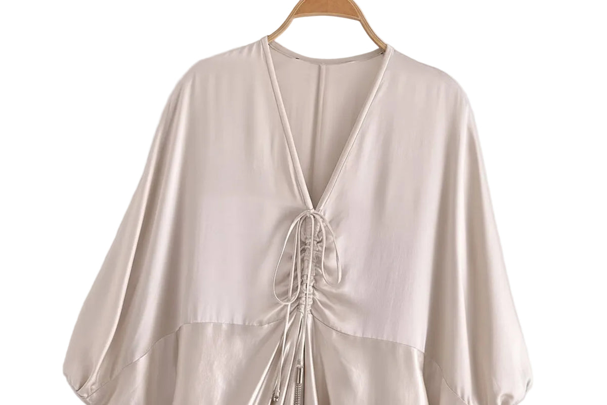 A light beige satin-like wrap top with a deep V-neckline and front lace-up detail, hanging on a wooden hanger against a plain light gray background.